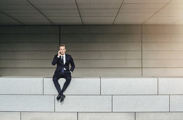 Businessman speaking phone stitting on brick wall