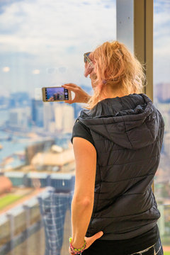 Aerial view of Hong Kong skyline. Observation deck with blurred background. Back of young tourist taking picture with smartphone enjoying view over Victoria Harbour. Travel and tourism asia concept.