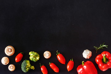 Stylish vegetarian background. Fresh vegetables and mushrooms over black board. Bell pepper, tomato, broccoli, champignon, garlic and tarragon. Copy space, top view.