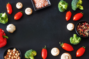 Vegetarian food frame over black background. Fresh vegetables and seasoning. Bell peppers, cherry tomatoes, broccoli, champignons, Himalayan salt, ground pepper and cardamom. Copy space.