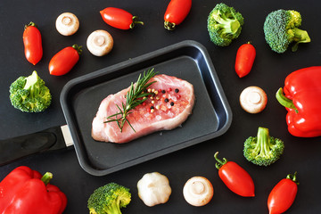 Raw pork or beef steak ready for frying or grill with fresh vegetables. Bell pepper, cherry tomato, broccoli, champignons over black background. Top view.