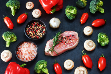 Raw veal or pork steak with seasoning and fresh vegetables ready for roasting or bbq. Bell pepper, broccoli, mushrooms, cherry tomato over black background. Top view.