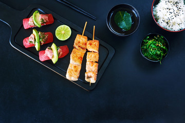 Top view of raw tuna sushi, grilled salmon on skewers, wakame, bowl of steamed rice, green tea over dark background with a copy space. 