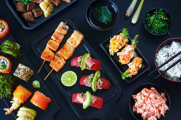 Top view of variety of japanese dishes over dark background. Tuna sushi, assorted sushi rolls on board, grilled salmon, sliced beef steak, wakame, crab meat, bowl of rice and cup of green tea.