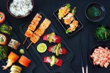 Top view of variety of japanese dishes over dark background. Tuna sushi, assorted sushi rolls with salmon, avocado, crab, bowl of rice, crab salad, wakame and green tea. 
