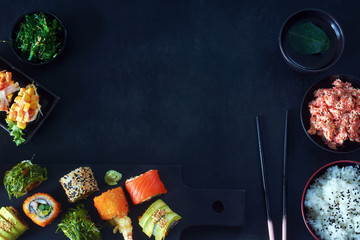 Top view of  assorted sushi rolls with salmon, avocado and wakame, crab meat, wakame salad, bowl of rice, green tea and chopsticks over black background. Copy space.