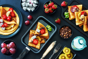 Top view of healthy breakfast set: thin pancakes, strawberries, exotic fruits, chocolate granola and milk over black background. 