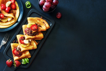 Thin pancakes with strawberries, grapes, kiwi, lime and honey over black background. Gourmet healthy breakfast with crepes and fruits. Copy space.