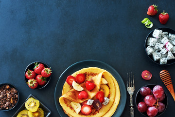 Crepes with chocolate granola, honey, strawberries, grapes, dragon fruit and golden kiwi. Top view of delicious thin pancakes with exotic fruits over dark board. 