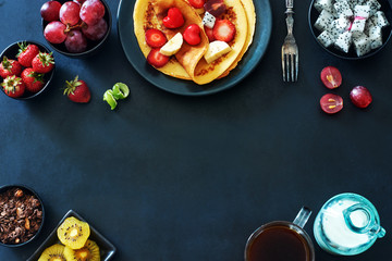 Obraz na płótnie Canvas Top view of breakfast with crepes with strawberries, grapes, kiwi, chocolate granola, coffee and milk over dark background. Food frame with space for text. 