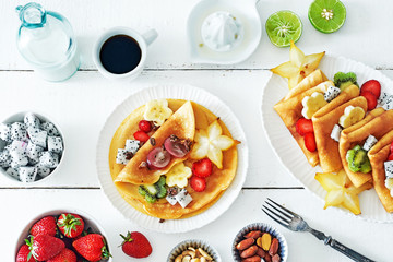 Healthy breakfast with thin pancakes, exotic fruits (starfruit, pitaya, banana, kiwi), strawberry, figs, granola, coffee and milk on a white table. 