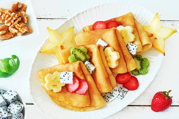 Close-up of thin pancakes with exotic fruits on a white table. Crepes with starfruit, banana, dragon fruit, kiwi and strawberry on a wooden table. 