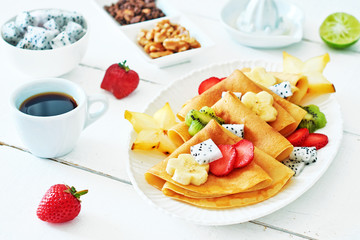 Close-up of thin pancakes with exotic fruits over white background. Crepes with starfruit, banana, dragon fruit, kiwi and strawberry, walnut, granola and coffee. 