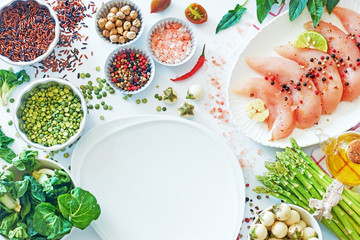 Top view of healthy dinner ingredients and white plate at the bottom. Raw chicken meat, dry brown rice and peas, fresh asparagus, eggplant and spinach, salt, pepper and olive oil.