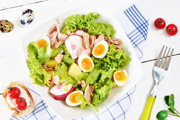 Top view of healthy green salad with lettuce, tuna, quail eggs, radish and potato and bruschetta with roasted tomato on a white table. 