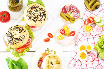 Top view of different bagels with ham, egg, salami, smoked salmon, veggies, cream cheese and pesto on white table. 