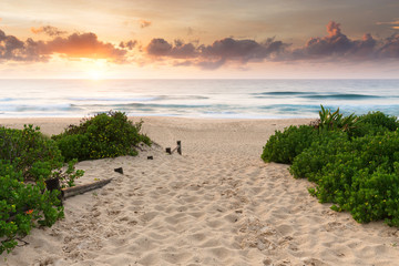 A colourful sunrise over a beautiful ocean scene and a walkway, spotted with footprints in the sand.