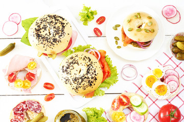 Different bagels with ham, salmon, salami, pickles, egg, lettuce, onion, radish, tomato on white table. Top view of healthy lunch.