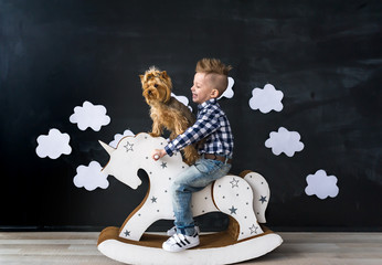 Cute baby boy riding wooden traditional rocking horse toy in room. The boy and yorkshire terrier.