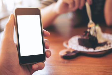 Mockup image of black mobile phone with blank white screen with brownie cake in ceramic plate on wooden table in background
