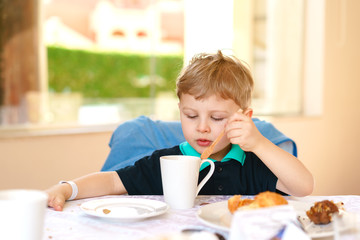 Obraz na płótnie Canvas Small blond boy enjoys his breakfast, sitting at a table in a resort cafe.