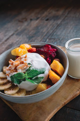 The plate of exotic fruits ( mango, banana, dragon fruit and papaya) and yogurt on top in the bowl on the wooden table with cocnut milk and spoon, Indonesia, Bali