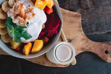 The plate of exotic fruits ( mango, banana, dragon fruit and papaya) and yogurt on top in the bowl on the wooden table with cocnut milk and spoon, Indonesia, Bali