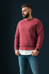 A young bearded handsome male hipster, dressed in a red pullover with long sleeves and jeans, stands indoors. - obrazy, fototapety, plakaty