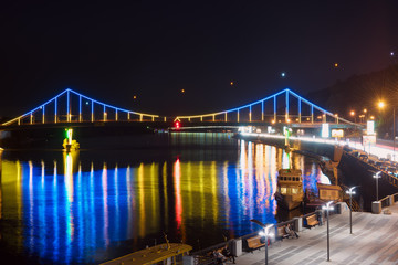 Night landscape. The city of Kiev, Ukraine, Europe. Pedestrian bridge across the Dnieper River. Beautiful lighting and reflection in water in blue and yellow colors