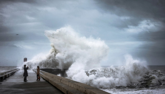 Crazy brave people on the pier near the lighthouse make photos during heavy storm against the backdrop of huge ocean waves. The Atlantic coast. Douro river mouth. Porto. Portugal. Europe.