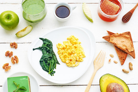 Healthy Green Meal On A White Table. Green Breakfast Or Lunch Concept: Scrambled Eggs With Spinach, Green Smoothie, Matcha Cake, Black Coffee, Brown Sugar, Peanut Butter Toasts, Apples. Top View.