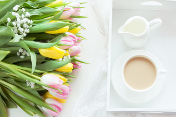 Morning breakfast in spring with a cup of black coffee with milk in the pastel colors, a bouquet of fresh yellow and pink tulips on a white background. Top view.