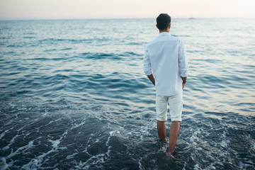 Horizontal back view of man wearing white shirt and pants on the beach and sea background. Travel vacation holiday. Man walking barefoot at the sea. Relax caucasian man model looking away, freedom. 