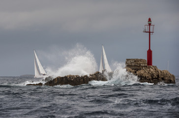 Two sailing boat in stormy weather close dangerous rock with red lighthouse
