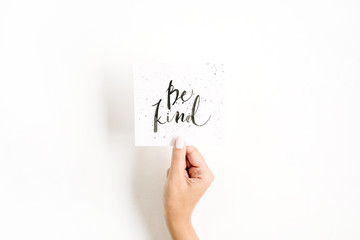 Minimal pale composition with girl's hand holding card with quote Be Kind written in calligraphic style on paper on white background. Flat lay, top view