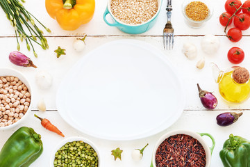 Brown, ruby and black rice in bowls, peas, garbanzo beans, assorted fresh vegetables, olive oil and spices. Empty plate in the middle. Vegetarian meal background. Copy space.