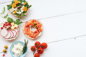 Variety of bagels with cream cheese, rocket salad, cherry tomatoes, italian ham, radish, salmon, olives, feta and tangerine on a wooden plate. Whole bagel on a white wooden table. Copy space.