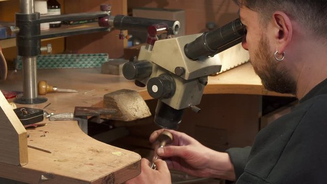 Young caucasian jeweler looking through the microscope. Bearded man with piercing in his ear instaling gemstone into the jewlery item. Male specialist using pusher to put the gem on the right place