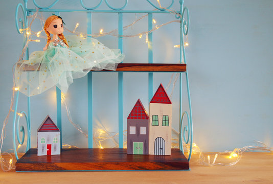 Cute doll and wooden little houses next to warm garland lights in front of blue background

