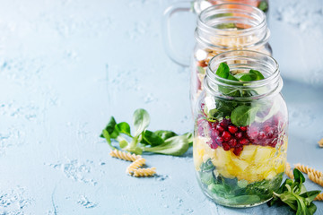 Variety of salads in mason jars. Fruit salad mango, pomegranate, greens, vegetables, wholegrain pasta, carrots and cauliflower, salmon. Standing over blue texture background. Food to go. With space