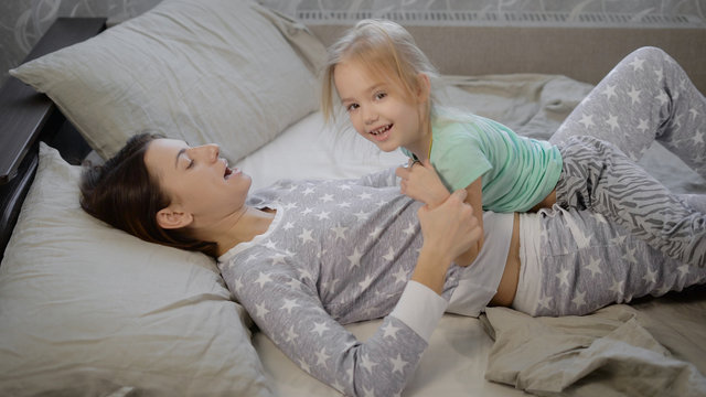 Young mother and her daughter lying in bed and fool around with each other in their weekend. Both girls are dressed in bright pajamas. Mom has short dark hair, his daughter blond, tied in a ponytail