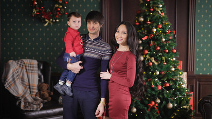 Father mother and their little son together looking into the camera on the background dressed Christmas tree. The boy turns around, looks around and shows a finger.