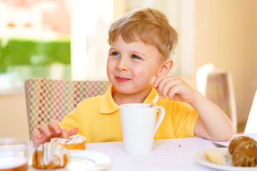 Happy kid sits at a table with desserts on a summer terrace lit with bright daylight. Boy looks away. He dressed in casual yellow polo.