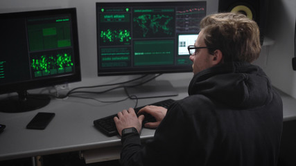 Experienced hacker carries out cyber attacks on major companies server to steal money from their accounts. Man with glasses at the same time work for multiple computers in an illegal secret office.