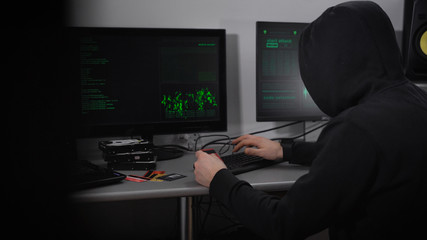 Back view of a hooded hacker making cyber attack on bank network. Man in black is typing fast on keyboard and stealing data of credit cards with the process viewed on screens in front of him.