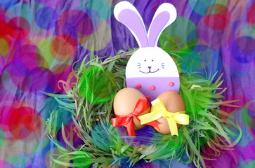 easter hand made decorated greeting card: yellow eggs and hand made festive plastic foam bunny in green grass twigs nest on purple background with inscription happy easter