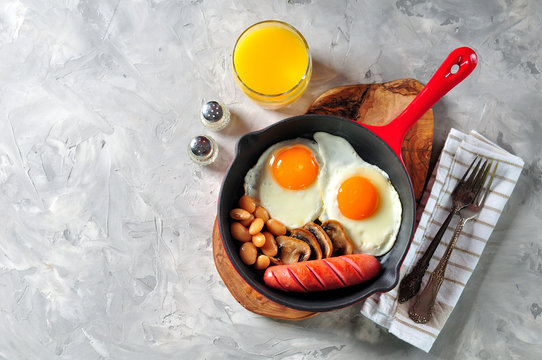 Classic breakfast of fried eggs, sausages, mushrooms, beans in a cast-iron frying pan.