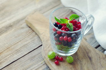 Fresh juicy berries of a gooseberry, black and red currant in a transparent glass mug on a wooden surface on a light background. Summer harvest of berries