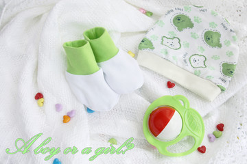 greeting card: green knitwear newborn baby booties and hat with colorful rattle on crocheted blanket white background with colorful hearts with with inscription a boy or a girl