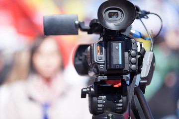 Video camera in the focus, blurred female reporter in the background. Television broadcasting.